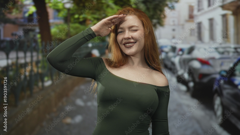 Fototapeta premium Redhead woman waving hand with bare shoulder visible in a green fitted dress on a cobblestone street lined with parked cars and a metal fence, smiling warmly; joy confidence.