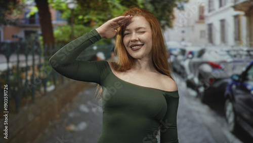 Quadro em tela Redhead woman waving hand with bare shoulder visible in a green fitted dress on a cobblestone street lined with parked cars and a metal fence, smiling warmly; joy confidence