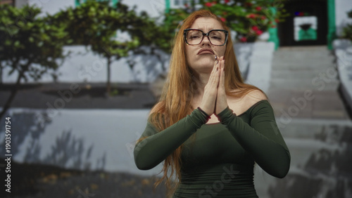 Schilderij op canvas Redhead woman presses hands together in pleading prayer at a white building entrance with steps and planters, glasses and green off shoulder top; pleading vulnerability