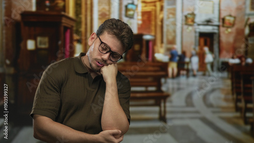 Man with glasses hand on chin and arm cradling elbow wearing olive polo seated in ornate church pews near altar; quiet contemplation.