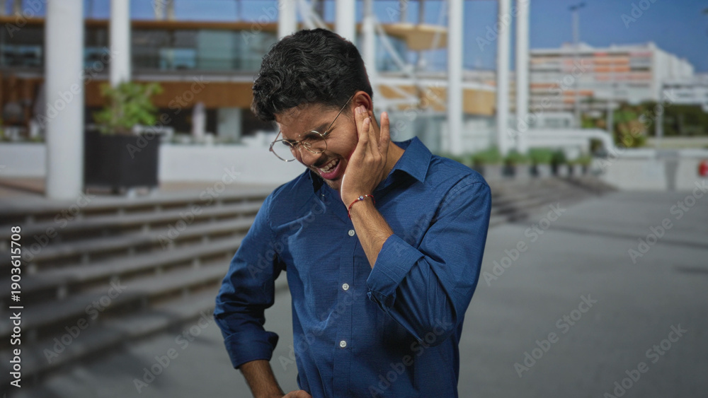 Fototapeta premium Man holding cheek with hand, wearing glasses and blue shirt, wincing near public building steps and plaza entrance; discomfort reflection.