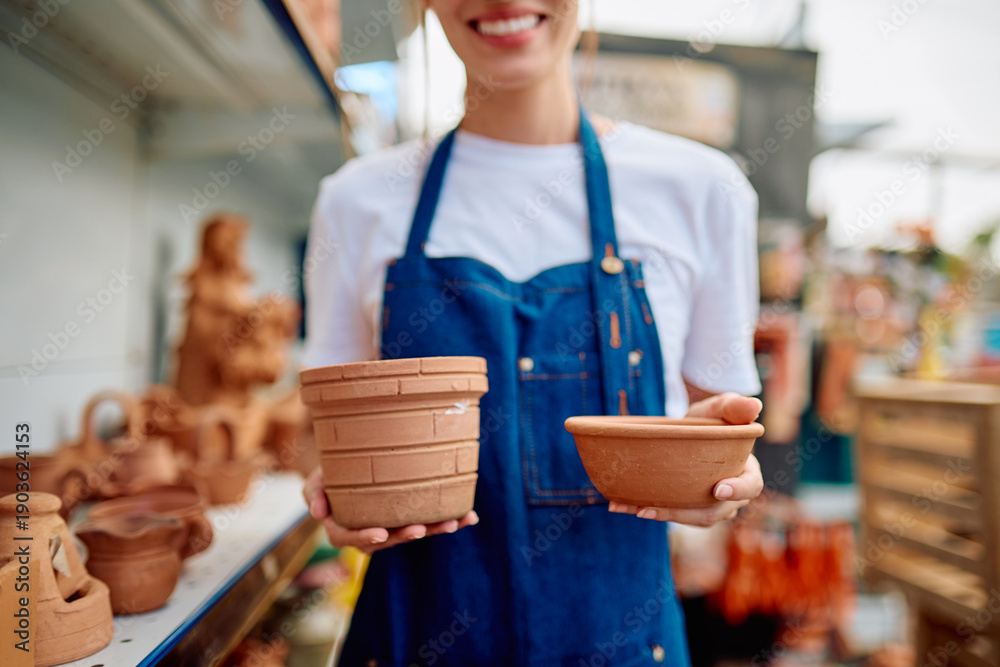 Fototapeta premium Woman artisan wearing an apron presenting two unglazed terracotta pots, offering traditional handmade ceramics at an outdoor market stall