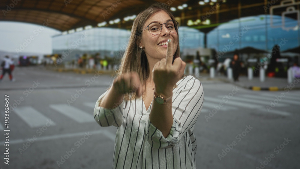 Fototapeta premium Blonde woman flashing middle finger and miming a crank motion outside an airport terminal arrivals area; defiance insult.