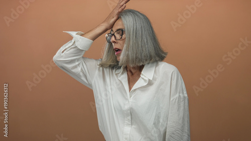 Woman with gray hair holds hand to forehead in studio with peach backdrop and white shirt; surprise realization.