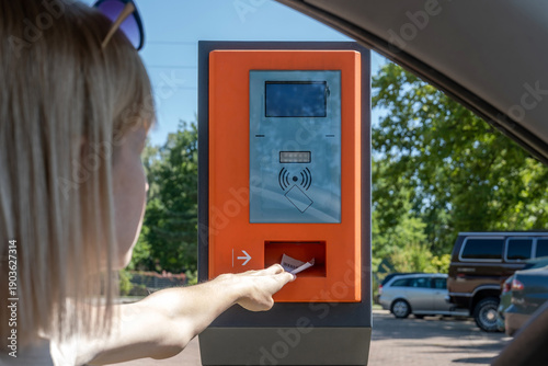 Woman reaching from car to parking ticket machine kiosk with blank screen and contactless symbol, self service payment or access control at entrance, sunny day, outdoor parking lot, copy space