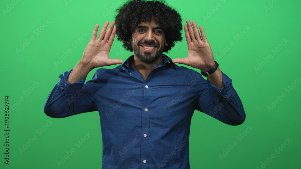 Fototapeta premium Man with hands up, palms facing camera and smiling, curly hair visible against a green studio backdrop; playful joy.