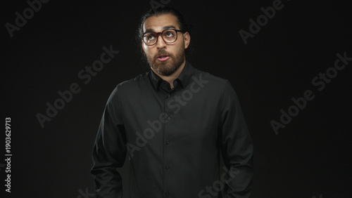 Man with glasses and beard, eyes closed and head tilted, hands in pockets wearing a dark shirt in a black studio; contemplative.