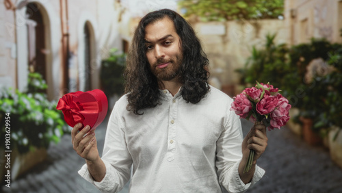 Fotografia Young man holds red heart box and pink bouquet while standing on a cobblestone street offering a gift; hesitant longing