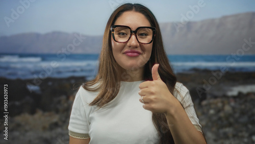 Bild auf Leinwand Woman adjusts tortoiseshell eyeglasses with hand to ear while gazing toward sunlit ocean waves on beach; listening inquiry attention curiosity