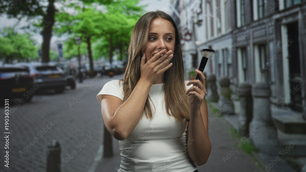 Fototapeta premium Young brunette woman holding makeup brush covers mouth with hand while smiling on a cobblestone city street lined with row houses; surprise playfulness.