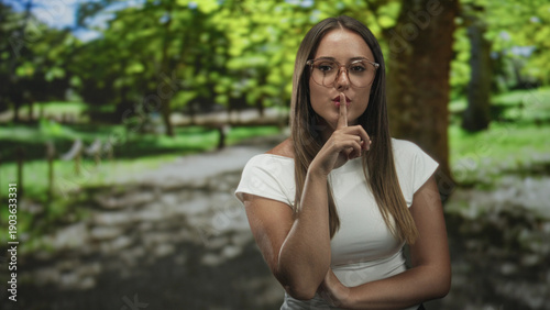 Картината върху платно Young hispanic brunette woman wearing glasses and a white top presses index finger to lips for silence in forest path; secret hush serenity