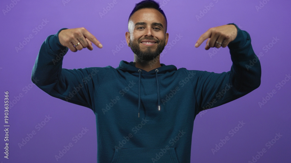 Naklejka premium Young hispanic man pointing down with hands in purple studio, wearing teal hoodie and smiling; promotion confidence.