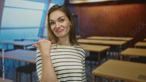 Photography Young caucasian woman pointing finger at unoccupied wooden desk in bright classroom building; contempt