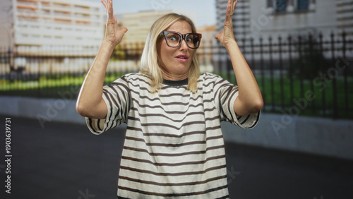 Schilderij op canvas Woman with glasses raises hands in shrug gesture on street in front of building and fence; surprise confusion