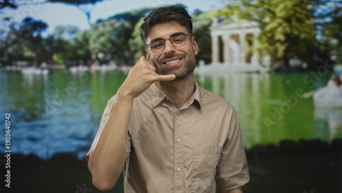 Young hispanic man wearing beige shirt making phone gesture with hand in green park; communication invitation friendly.
