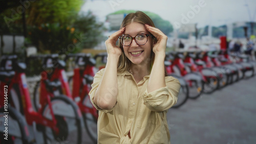Billede på lærred Woman wearing glasses presses temples beside red rental bikes lined in a row on busy city street; relief calm
