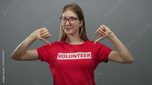 Foto Woman in red volunteer shirt standing confidently with thumbs pointing to chest in a grey studio; pride