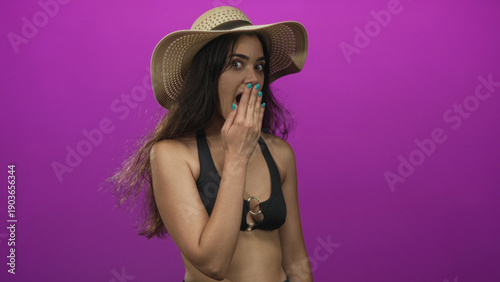 Fotografie Woman wearing black bikini top and straw hat with hand covering mouth laughing in purple studio; joy