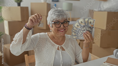 Fotografie Woman holding house keys aloft and fanning a stack of us dollar bills, smiling toward camera amid cardboard moving boxes inside building; home sale celebration