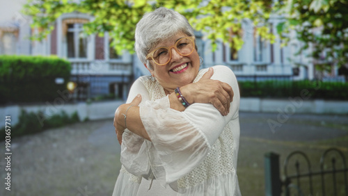 Tableau sur toile Woman hugging herself with hands crossed over chest in front of building, smiling broadly, wearing glasses, bracelet and white dress in a courtyard; self love joy