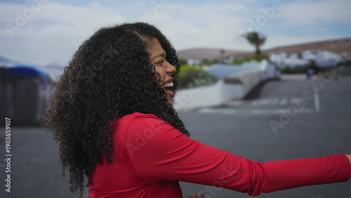 Fotografie African american woman in red shirt pulls an imaginary rope with her hand on a sunlit street outdoors; effort determination