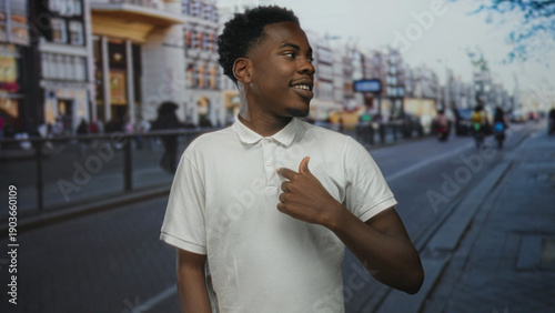 Fotografi Man pointing finger to white polo shirt against blurred sidewalk and storefront building facades in busy city street; pride