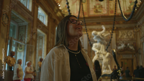 Cuadro en lienzo Woman looking up with visible face and glasses in ornate building gallery, wearing layered necklace and white shirt over black top; awe curiosity