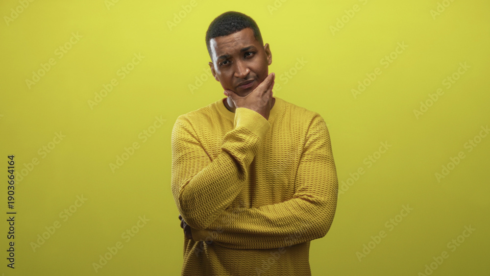 Obraz premium Young black man hand on chin in yellow sweater thinking pose in studio; introspection contemplation calm.
