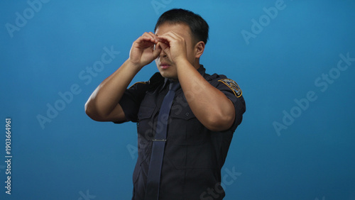 Fotografi Man wearing police uniform uses hand binoculars gesture over eyes in a steady posture against a blue studio wall; duty