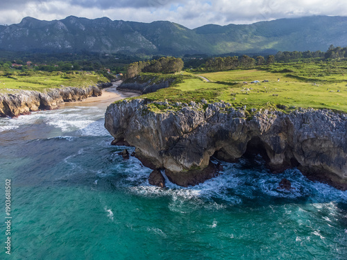 Wide-angle ocean view toward Playa de Guadamía featuring high rocky cliffs with sea caves, vibrant green meadows, and distant mountain peaks. Llanes, Asturias, Spain.