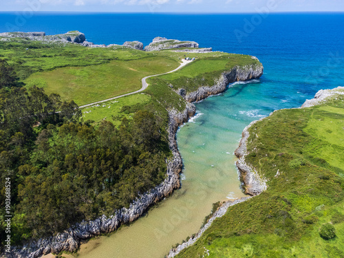 Aerial perspective of Playa de Guadamía, a narrow sandy beach carved deep into the limestone rock with calm, crystal-clear water. Located on the border between Llanes and Ribadesella, Asturias, Spain.