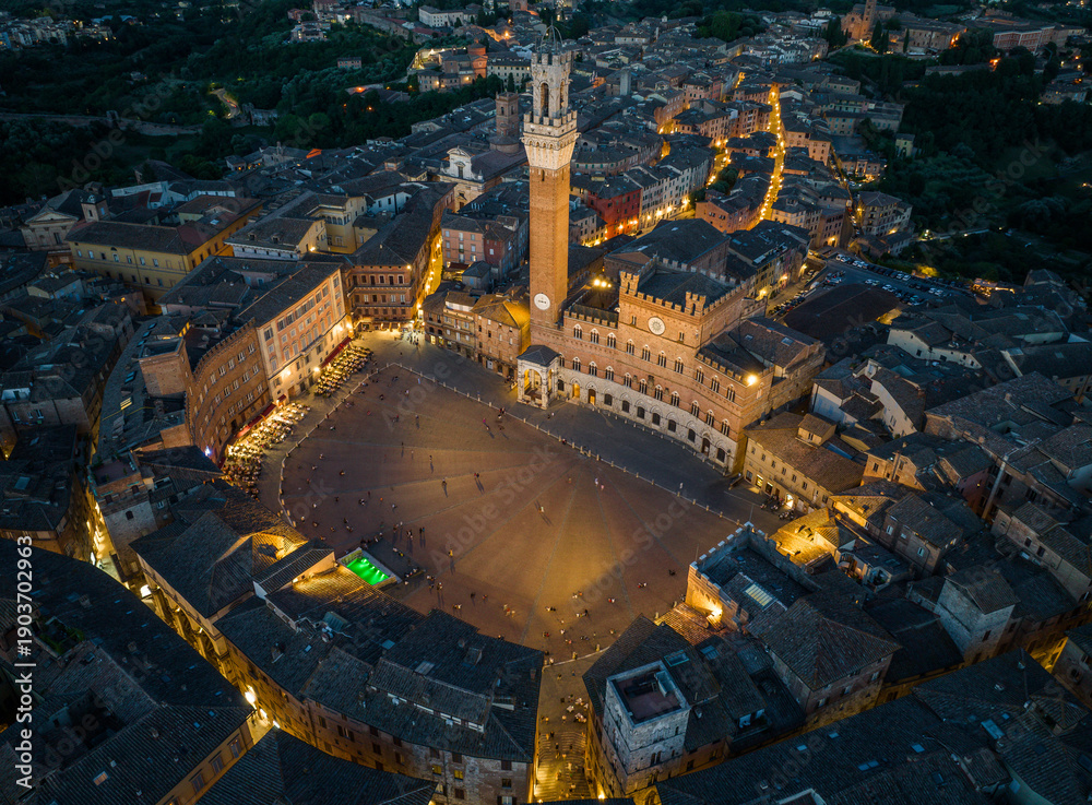 Fototapeta premium Aerial night view of Torre del Mangia rising above Piazza del Campo in Siena, Tuscany. Illuminated medieval tower and historic city center seen from above during evening hours