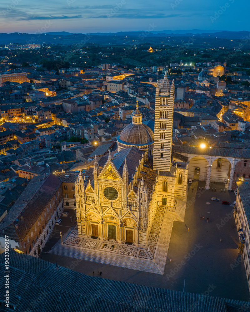 Fototapeta premium Aerial view of Siena Cathedral (Duomo di Siena) illuminated at blue hour, showcasing Gothic architecture and historic landmarks in the medieval city of Siena, Tuscany, Italy