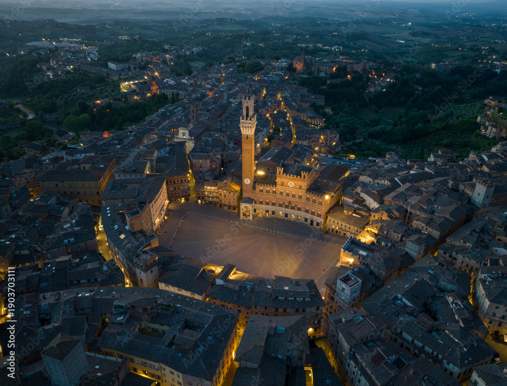 Fototapeta premium Aerial night view of Torre del Mangia rising above Piazza del Campo in Siena, Tuscany. Illuminated medieval tower and historic city center seen from above during evening hours