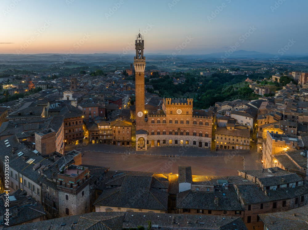 Fototapeta premium Aerial night view of Torre del Mangia rising above Piazza del Campo in Siena, Tuscany. Illuminated medieval tower and historic city center seen from above during evening hours