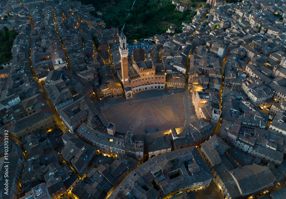 Fototapeta premium Aerial night view of Torre del Mangia rising above Piazza del Campo in Siena, Tuscany. Illuminated medieval tower and historic city center seen from above during evening hours