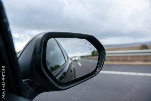A car's rear view mirror shows a blurry reflection of a car in the distance