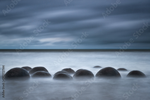 Fotografie Long exposure seascape featuring the Moeraki Boulders in New Zealand under a moody, overcast sky with misty water effects