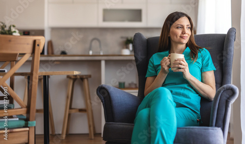 ng adult female healthcare professional wearing scrubs and an id badge, smiling while holding a cup of coffee and looking out a window during a relaxing moment in a contemporary kitchen.