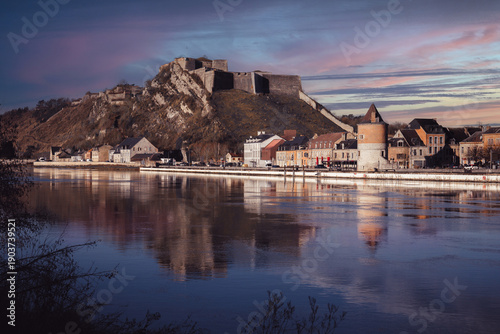 La ville de Givet au coucher du soleil, sur les quais de la Meuse avec le Fort de Charlemont, la tour Victoire et les habitations au bord de l'eau.