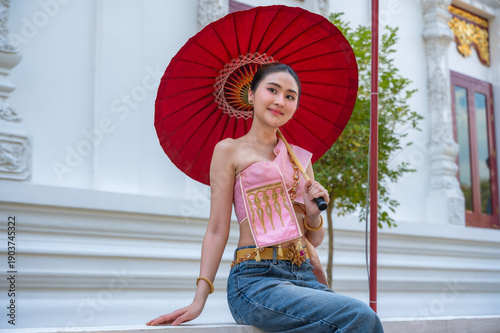 Thai woman in contemporary trendy Sabai mix and match with jeans holding red umbrella popular street fashion in Thailand sitting in temple
