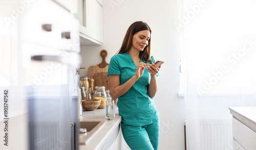 A healthcare worker in scrubs enjoys a moment of relaxation at home, checking her smartphone and holding a glass. The setting is a modern kitchen filled with natural light.