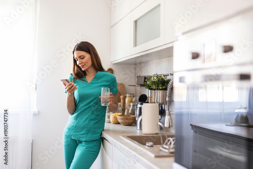 A healthcare worker in scrubs enjoys a moment of relaxation at home, checking her smartphone and holding a glass. The setting is a modern kitchen filled with natural light.
