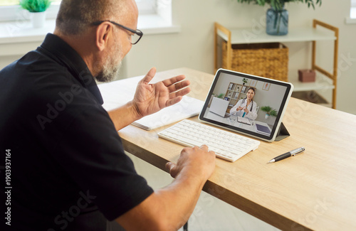 Fotografi Man on tablet consults doctor via telemedicine during video call at home