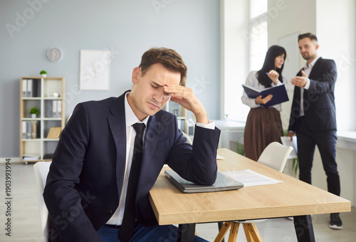 Tired man sits at office desk resting head in hand as colleagues talk in background. Scene reflects stress, anxiety, burnout, and workplace gossip. Man feels isolated and emotionally exhausted.
