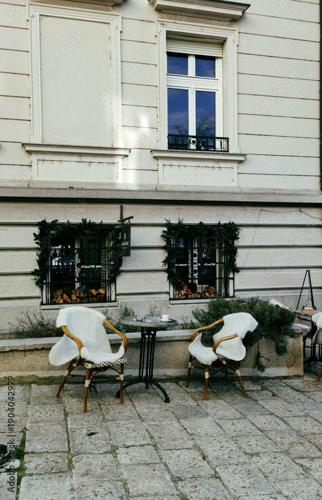 Obraz premium Cozy outdoor cafe terrace with wicker chairs and small table near classic building facade. Calm European atmosphere with vintage tones and subtle film grain