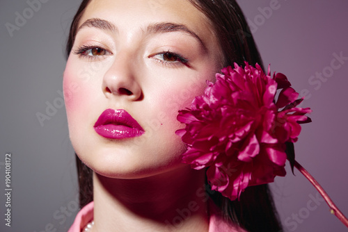 Close-up of woman with glossy bright pink lips and smooth skin holding a vivid pink flower near her face. High-fashion beauty portrait on grey background symbolizing elegance.