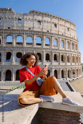 A young woman beams with joy while using her smartphone at the iconic Colosseum, a monumental structure in Rome, fully embracing the lively atmosphere that surrounds this historic landmark