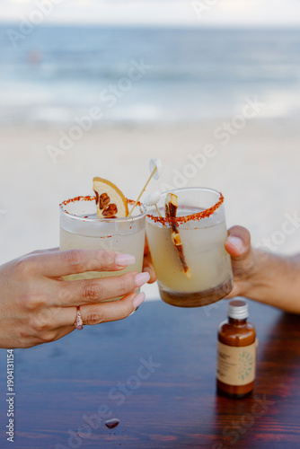 Two friends joyously clink their glasses filled with colorful and refreshing cocktails as they relax by the beautiful beach, perfectly capturing the essence of summer and the warmth of friendship