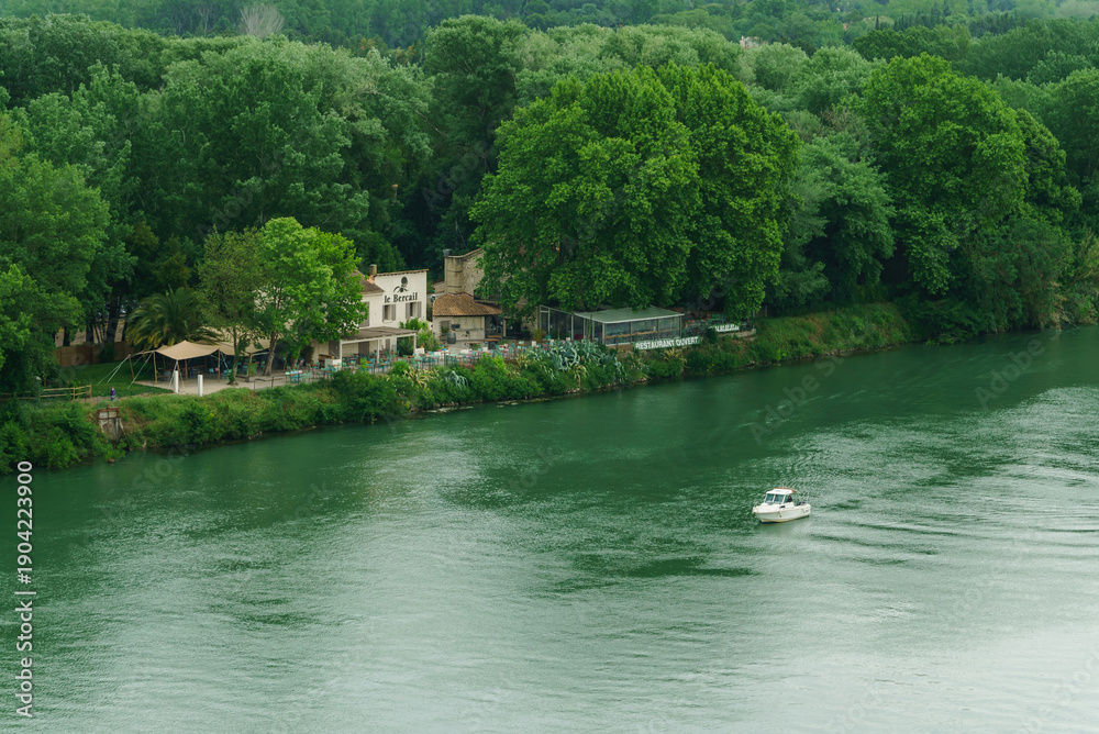 Obraz premium A small boat creates ripples on a calm river, surrounded by lush greenery and cozy cabins